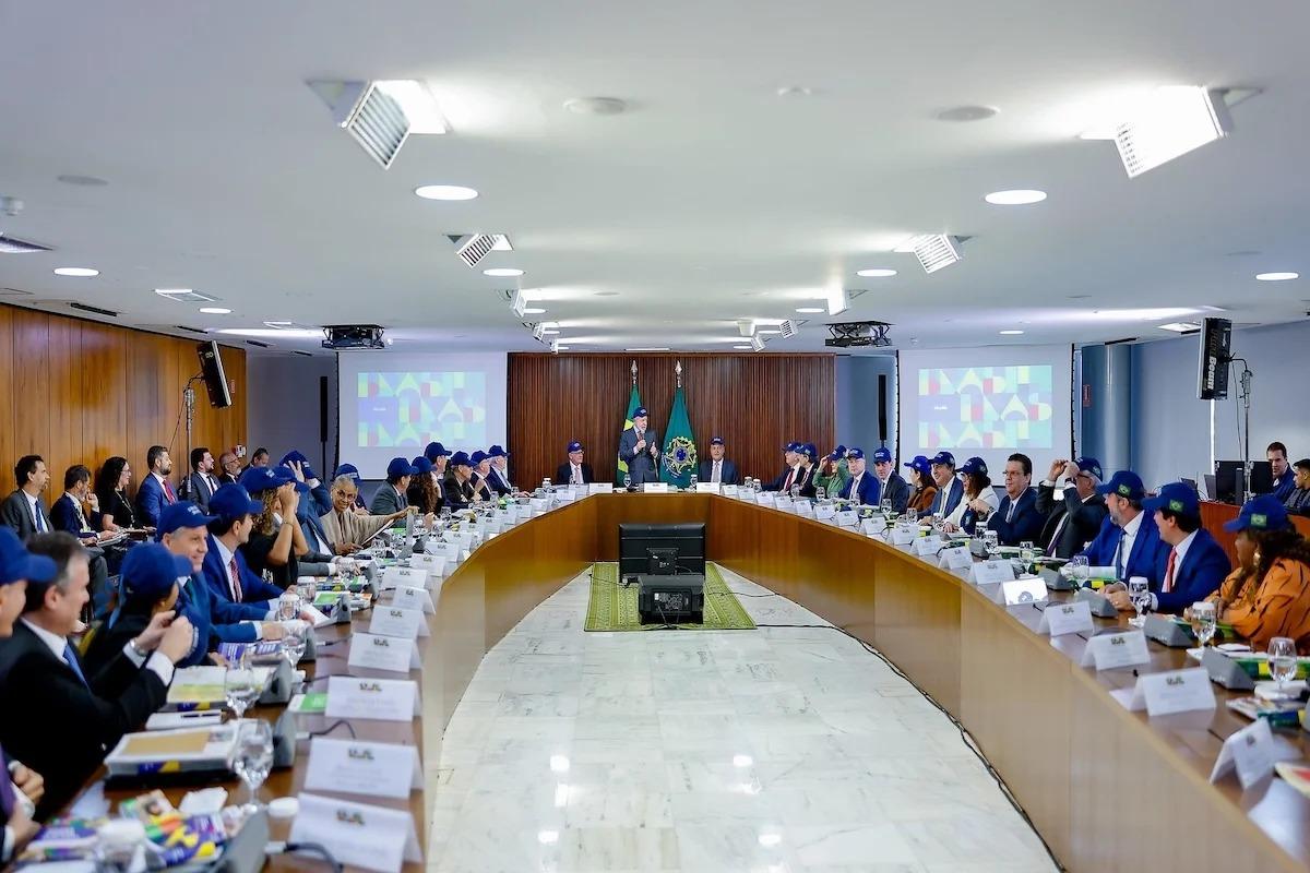 Reunião em uma grande mesa oval no Palácio do Planalto, com dezenas de participantes, muitos usando bonés azuis com a bandeira do Brasil. No centro da mesa, ao fundo, há duas bandeiras — do Brasil e da República Federativa do Brasil. Telões laterais exibem imagens coloridas com formas geométricas. A pessoa ao centro fala em pé, enquanto os demais acompanham sentados.