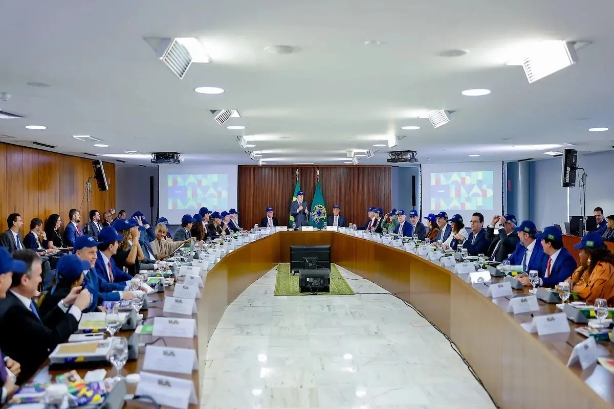 Reunião em uma grande mesa oval no Palácio do Planalto, com dezenas de participantes, muitos usando bonés azuis com a bandeira do Brasil. No centro da mesa, ao fundo, há duas bandeiras — do Brasil e da República Federativa do Brasil. Telões laterais exibem imagens coloridas com formas geométricas. A pessoa ao centro fala em pé, enquanto os demais acompanham sentados.