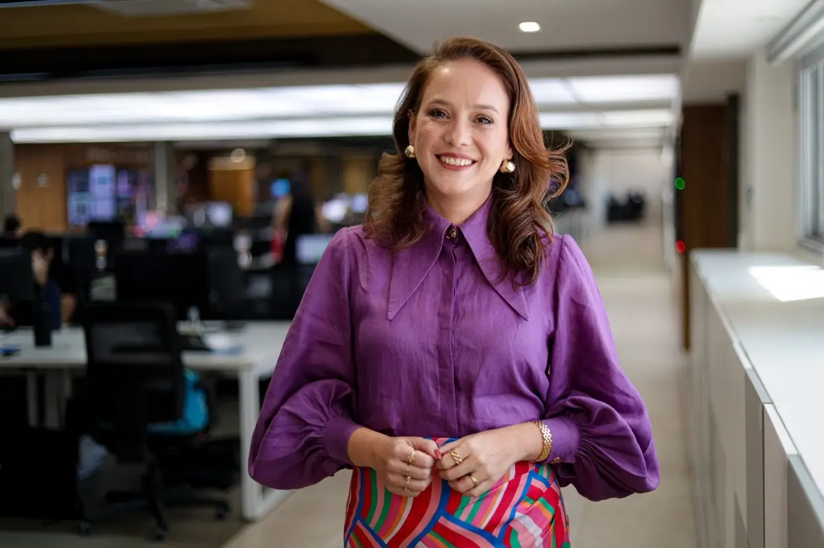 Mulher em pé, sorrindo levemente, usa camisa roxa de mangas bufantes e saia colorida com listras geométricas. Ela está em um ambiente de escritório, com mesas, cadeiras e pessoas trabalhando ao fundo.
