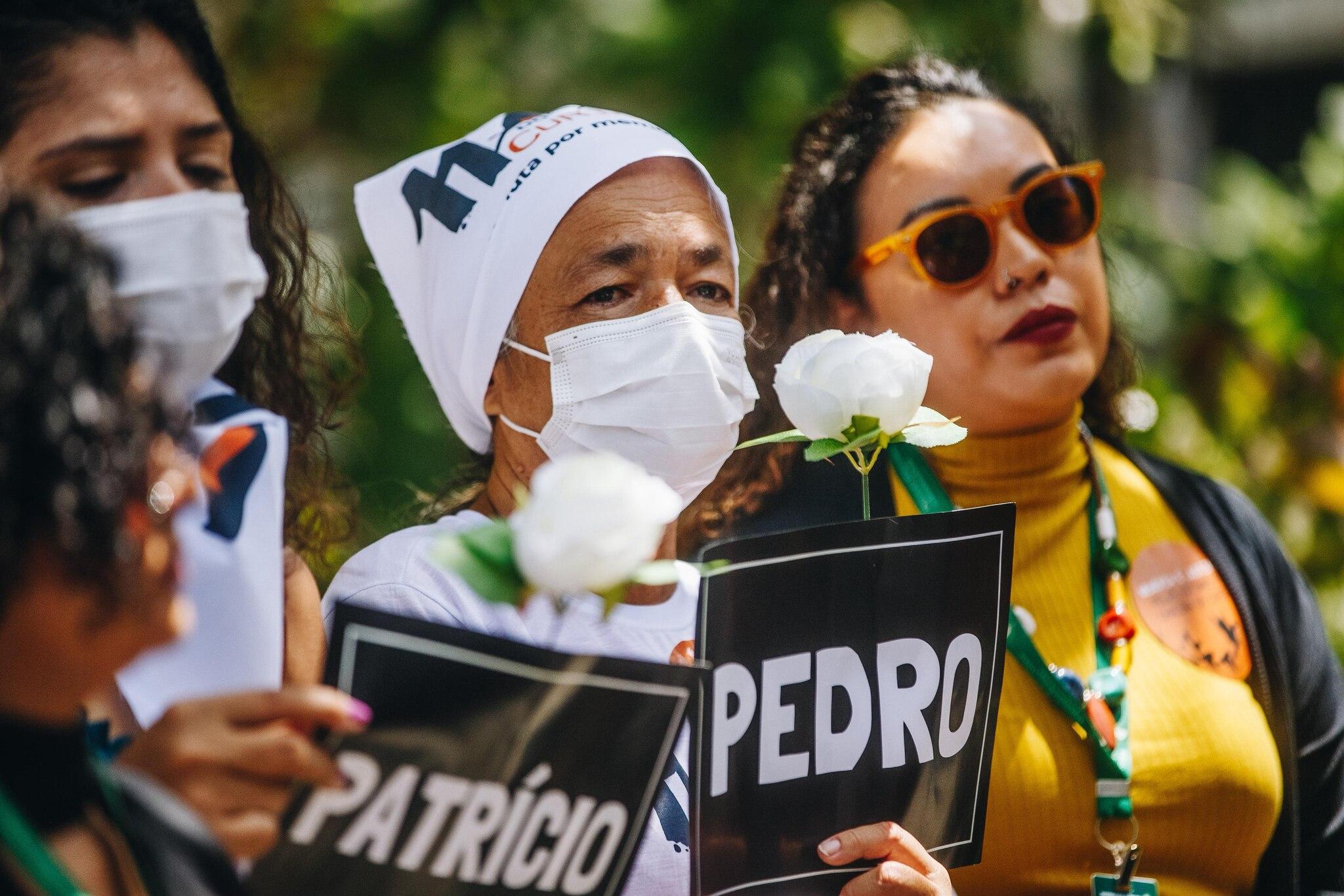 Mulher negra usa bandana na cabeça em alusão ao movimento Mães do Curió, segura flor branca e uma placa em que está escrito o nome Pedro