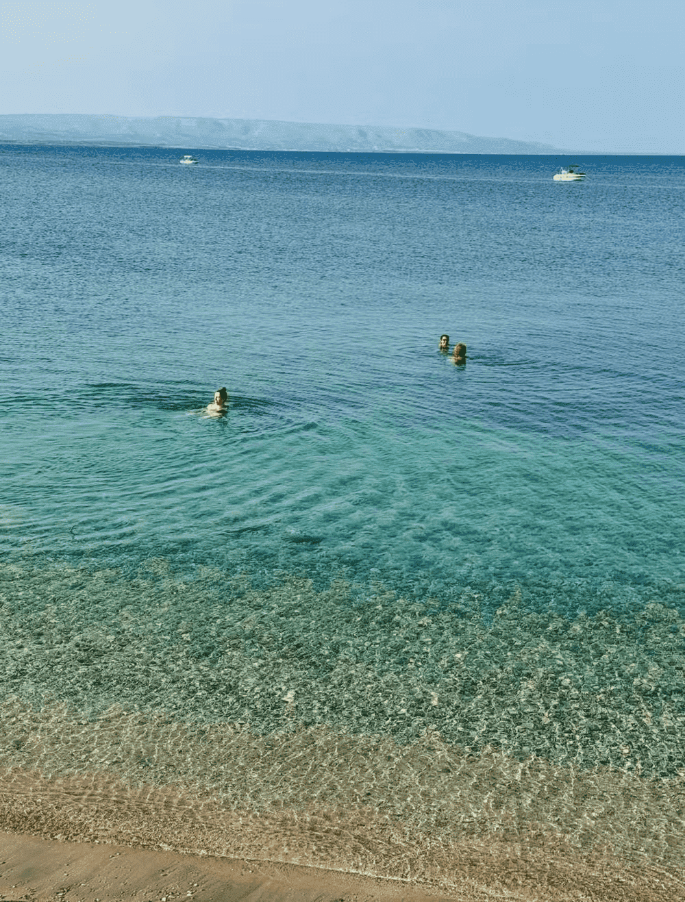 Praia de águas cristalinas com fundo de pedrinhas visíveis e faixa de areia clara em primeiro plano. Três pessoas nadam no mar calmo e azul, e ao fundo é possível ver dois barcos e uma faixa de terra montanhosa no horizonte sob um céu claro.