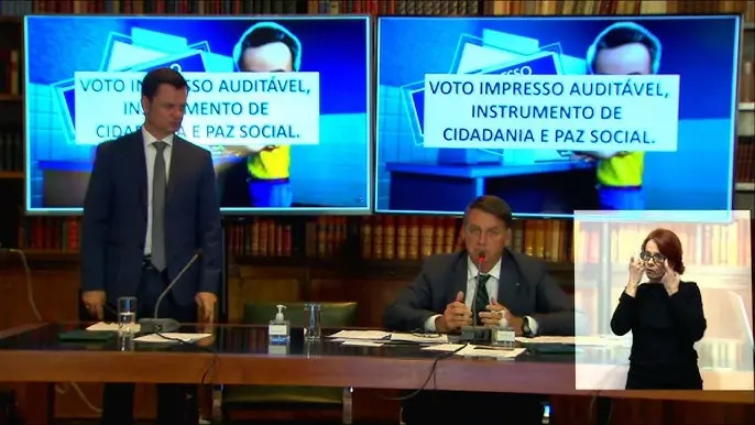 Bolsonaro sentado em uma mesa com duas TV exibindo a defesa do voto impresso
