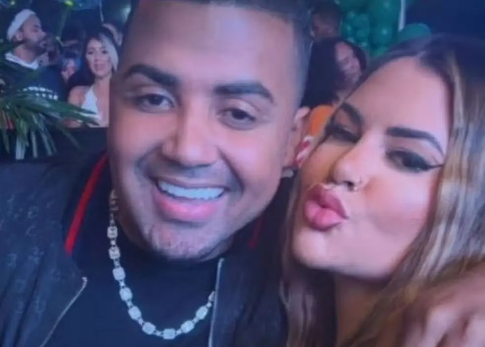 Imagem de um homem sorridente e uma mulher fazendo bico, em ambiente de festa com várias pessoas ao fundo, celebrando momentos de alegria.