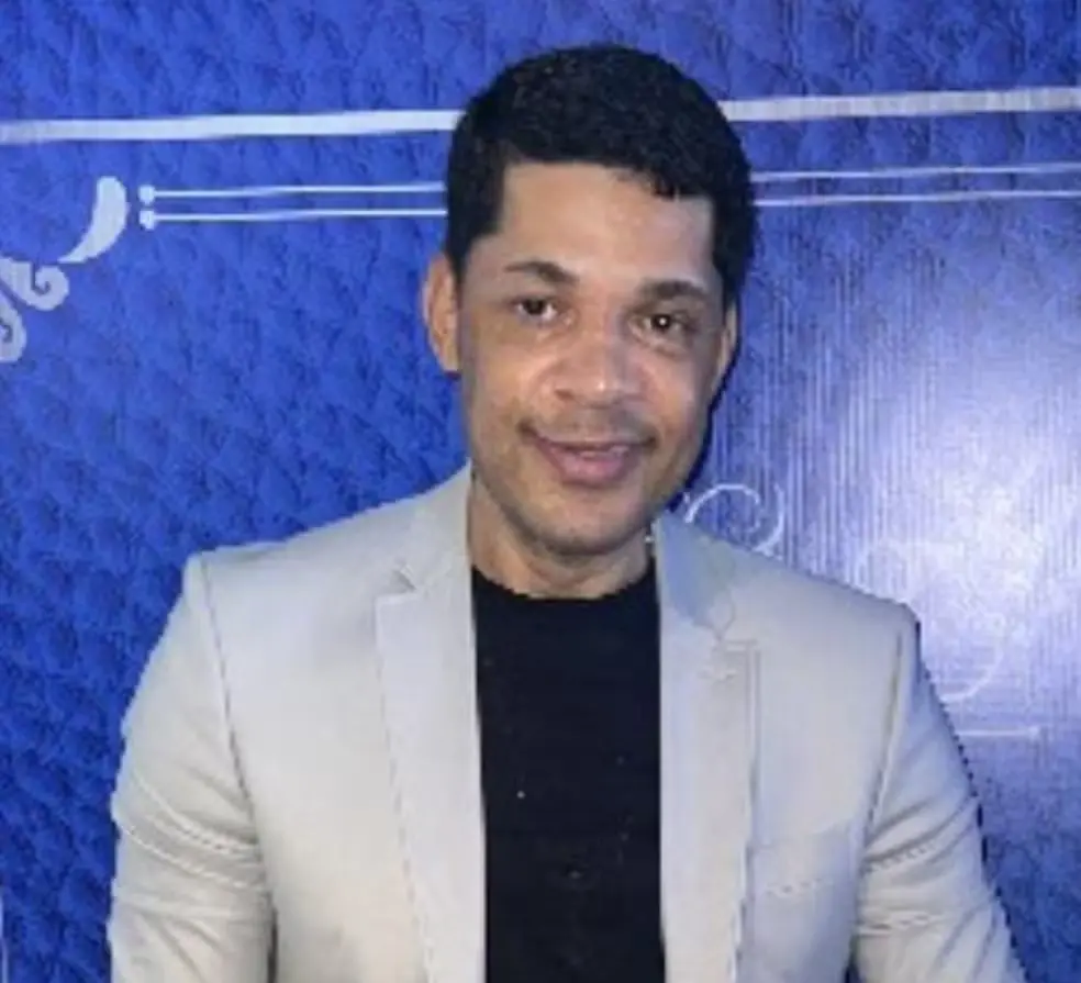 Homem sorridente com cabelo escuro, pele clara e vestindo terno cinza, sentado diante de uma parede azul com detalhes brancos, em ambiente de evento ou celebração.