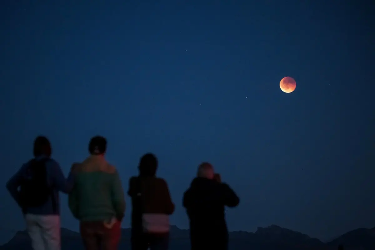 Quatro pessoas assistindo ao eclipse lunar sob um céu noturno com a lua parcialmente avermelhada, em uma paisagem de montanhas ao fundo.