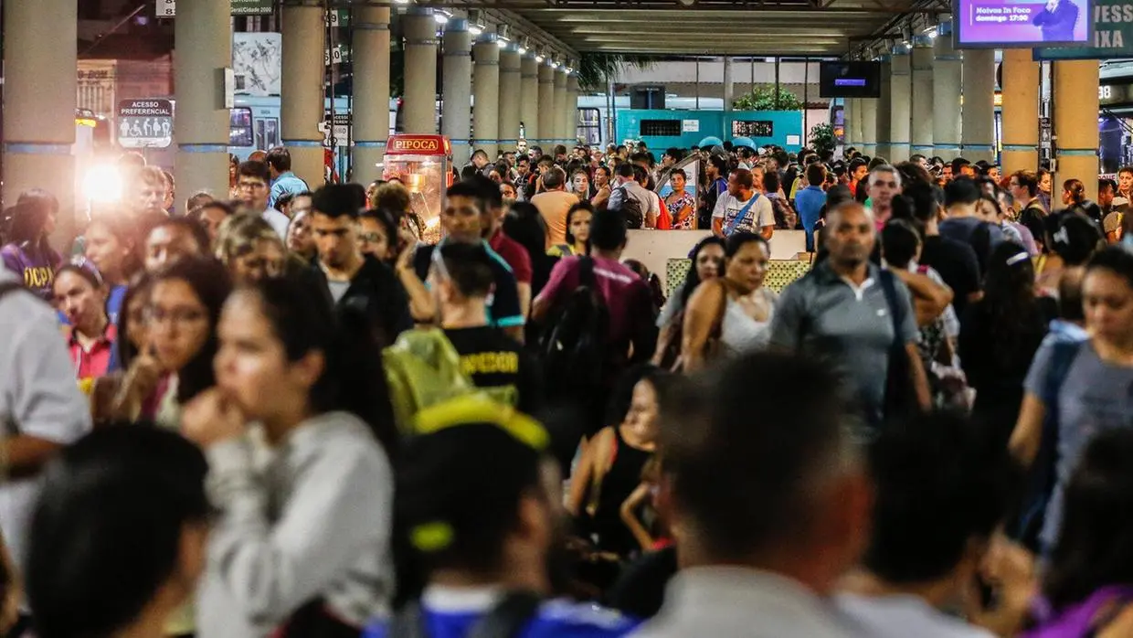 Grande aglomeração de pessoas em um terminal ou estação de transporte público. A multidão ocupa todo o espaço, com pessoas caminhando, esperando e conversando. Ao fundo, há uma máquina de pipoca iluminada e ônibus estacionados. O ambiente está movimentado e lotado.