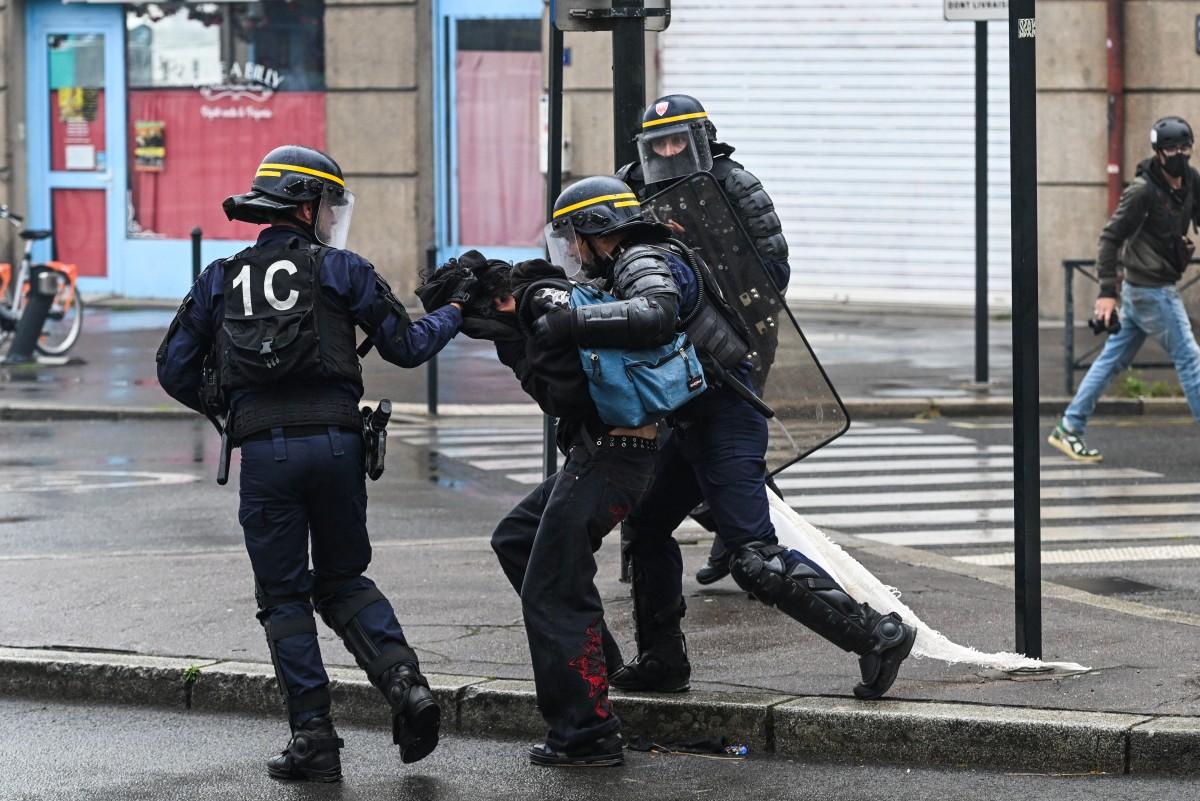 Policiais de face coletivo na rua, com equipamentos de proteção, detendo uma pessoa durante manifestação em Paris.