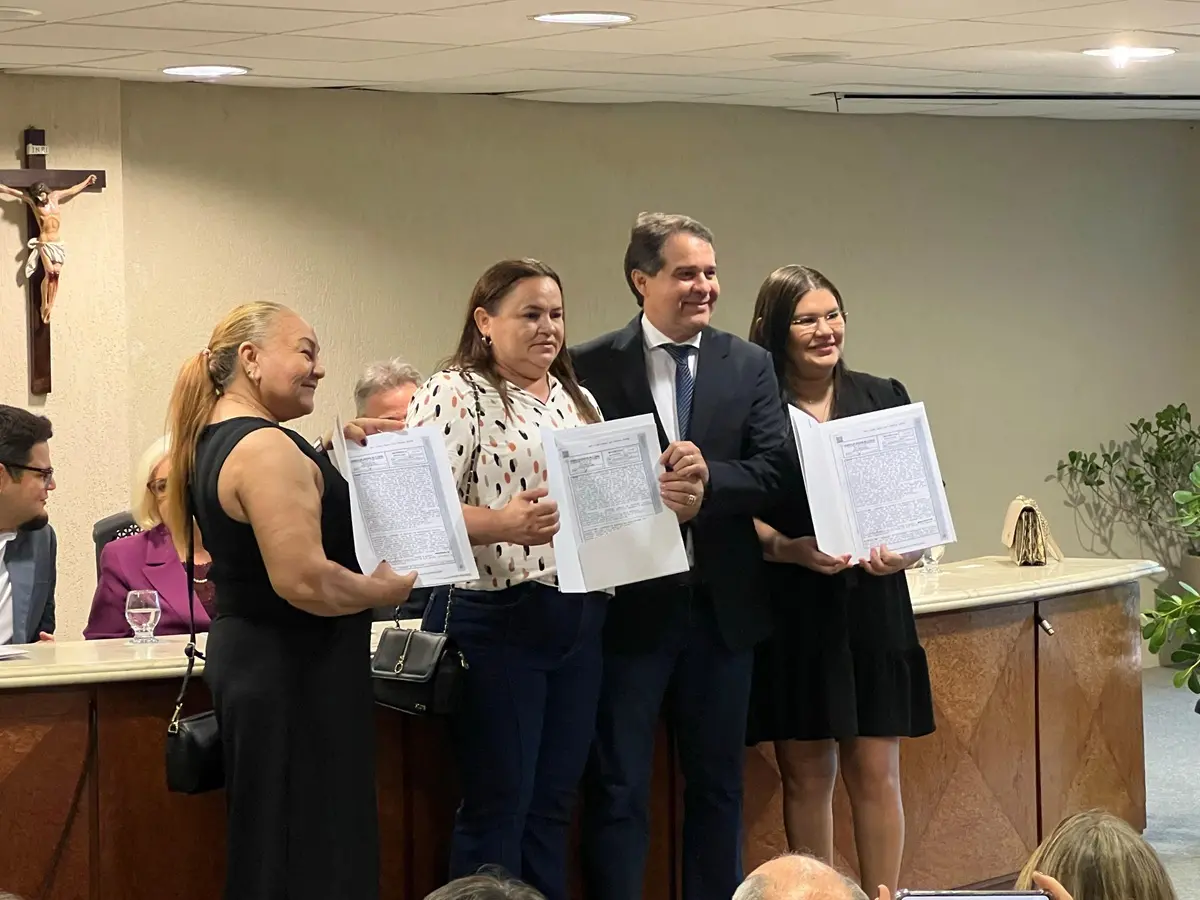 Três mulheres e um homem posam sorridentes em pé, segurando documentos de matrícula de imóvel durante cerimônia oficial.