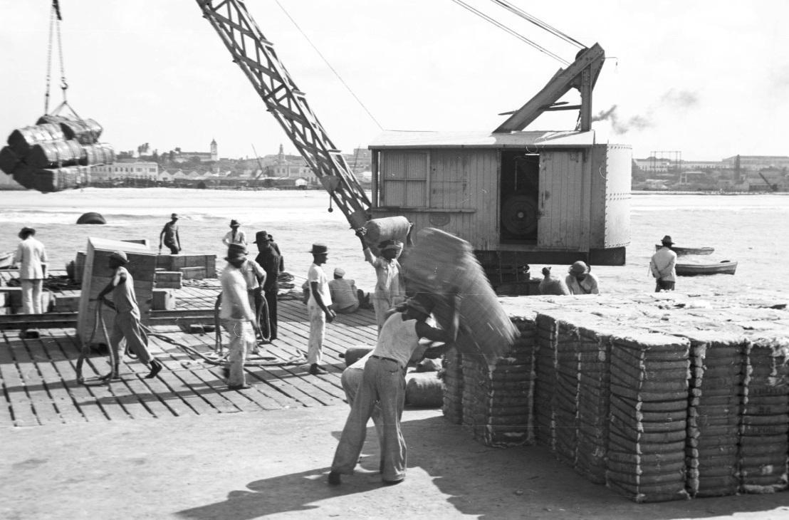 Uma fotografia em preto e branco de trabalhadores portuários no antigo Porto de Fortaleza. Um guindaste à esquerda está içando uma carga de fardos, enquanto homens descarregam e empilham sacos ou fardos semelhantes à direita. Outros trabalhadores estão no cais, observando o trabalho. Ao fundo, é possível ver o mar, barcos e a silhueta da cidade.