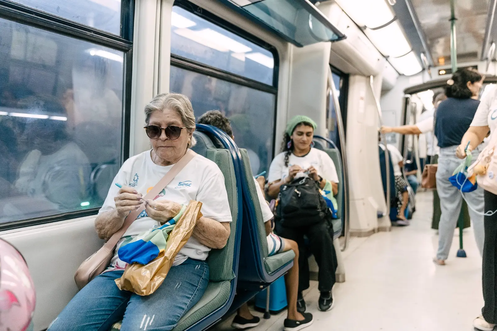 Mulher sentada no metrô, concentrada em fazer crochê, enquanto outra participante, mais ao fundo, também tricota; passageiros seguem em pé no corredor
