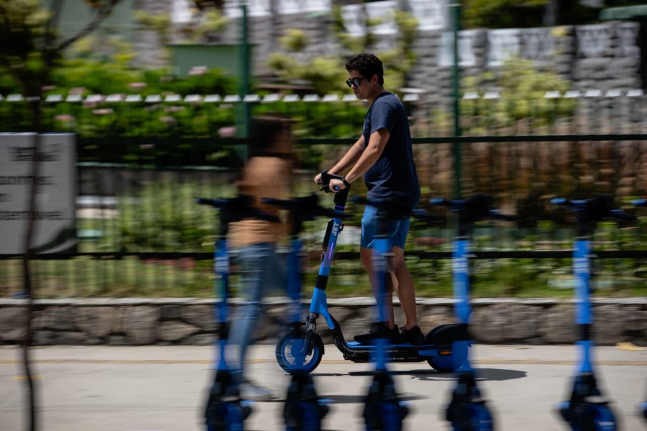 Rapaz jovem de blusa azul escuro usando capacete e óculos, andando de patinete elétrico na calçada, com uma pessoa em frame borrado ao fundo, em ambiente urbano com piso de cimento.
