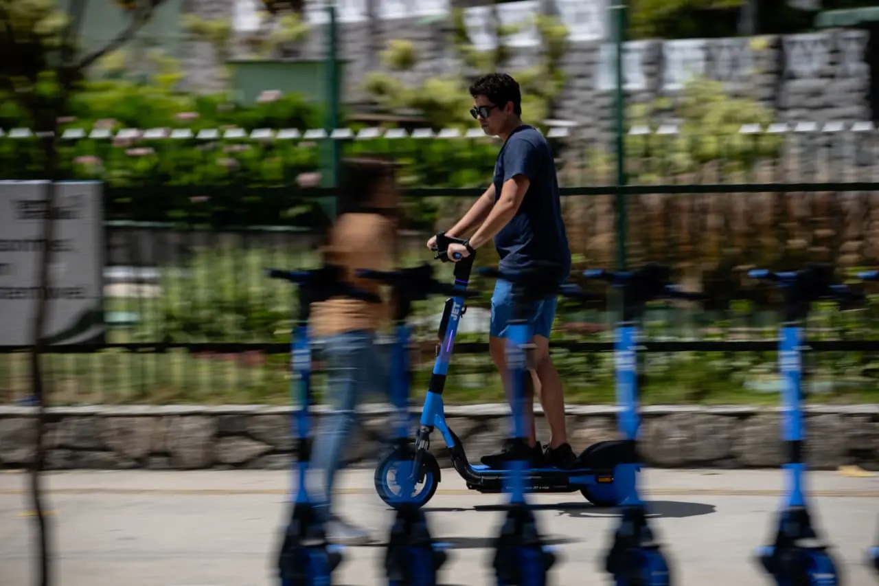 Rapaz jovem de blusa azul escuro usando capacete e óculos, andando de patinete elétrico na calçada, com uma pessoa em frame borrado ao fundo, em ambiente urbano com piso de cimento.