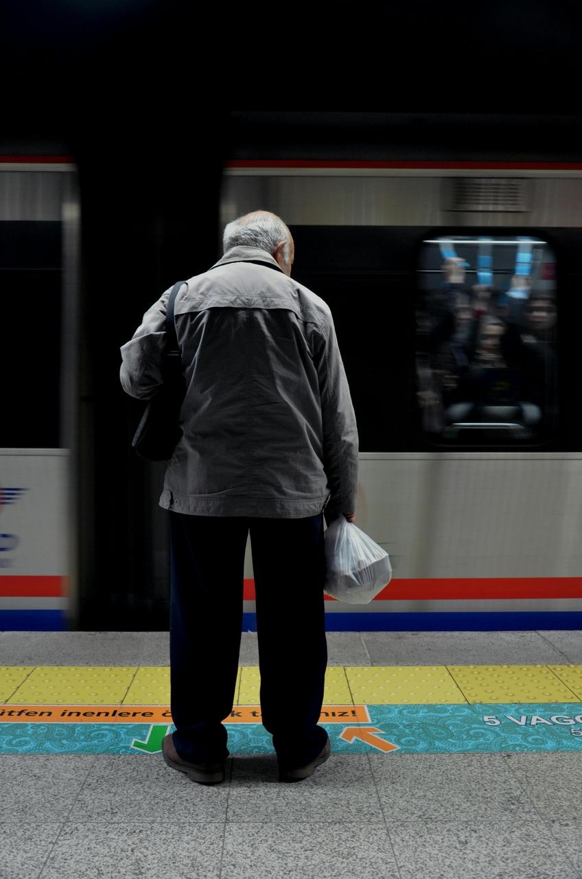 Pessoa idosa vista de costas, em pé na plataforma do metrô, segurando uma sacola plástica, enquanto um trem passa ao fundo