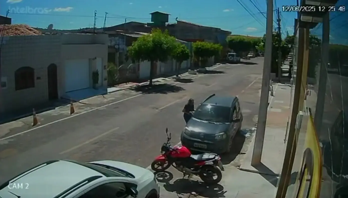 Imagem de câmera de segurança mostrando uma rua residencial durante o dia, com carros estacionados, uma motocicleta vermelha à frente e uma pessoa ao lado de um carro cinza.