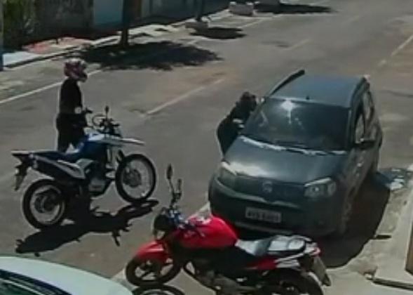Imagem de câmera de segurança mostra uma pessoa de capacete e armada em uma motocicleta parada ao lado de um carro cinza, enquanto outra pessoa interage junto à porta do veículo; uma moto vermelha está estacionada em frente.