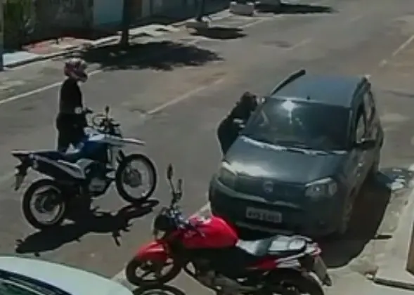 Imagem de câmera de segurança mostra uma pessoa de capacete e armada em uma motocicleta parada ao lado de um carro cinza, enquanto outra pessoa interage junto à porta do veículo; uma moto vermelha está estacionada em frente.