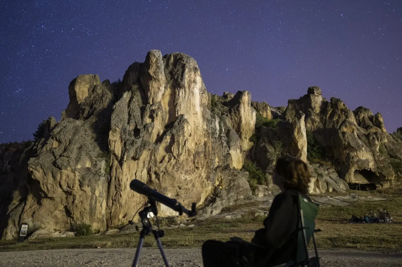 Pessoa observando o céu estrelado ao lado de uma grande formação rochosa, com um telescópio, durante a noite.