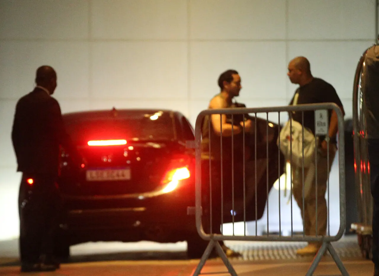 Foto desfocada de Allan saindo de carro preto com um segurança ao lado e uma silhueta de outra pessoa à esquerda, em fachada de hotel.