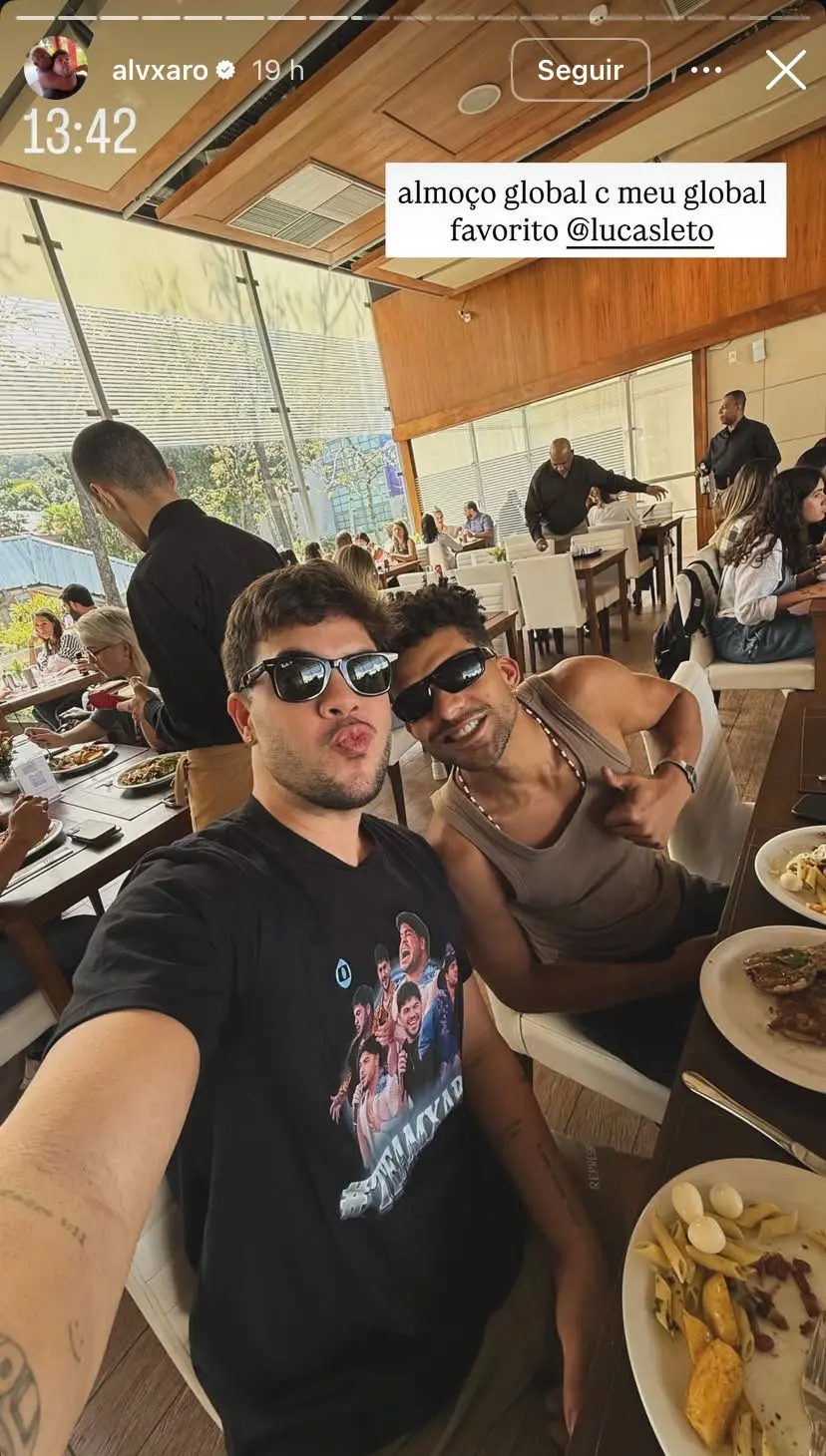 Alvaro e Lucas Leto sorrindo e curtindo almoço em restaurante sofisticado, com prato de comida na mesa, ambiente aberto com grande janela e decoração moderna, pessoas ao fundo