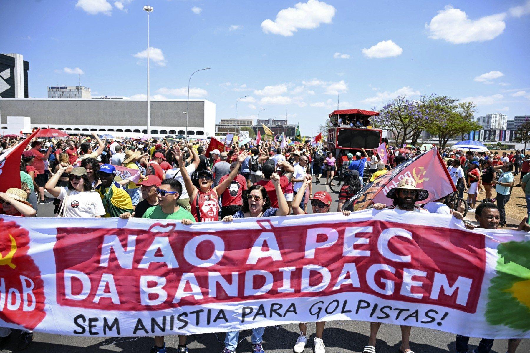 Manifestantes ocupam uma rua durante um protesto em Brasília, segurando uma grande faixa que diz: “NÃO À PEC DA BANDIDAGEM — SEM ANISTIA PARA GOLPISTAS!”. As pessoas estão vestidas com roupas de cores vibrantes, muitas usando bonés, camisetas com mensagens políticas e levantando os punhos em sinal de protesto. Há bandeiras de movimentos sociais e partidos políticos, incluindo o símbolo do Partido Comunista do Brasil (PCdoB). Ao fundo, um carro de som vermelho está cercado por mais manifestantes. O céu está claro e azul, indicando um dia ensolarado.