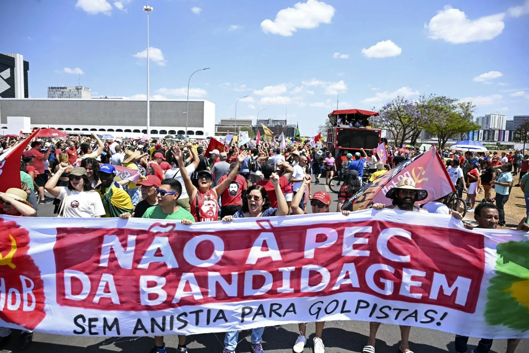 Manifestantes ocupam uma rua durante um protesto em Brasília, segurando uma grande faixa que diz: “NÃO À PEC DA BANDIDAGEM — SEM ANISTIA PARA GOLPISTAS!”. As pessoas estão vestidas com roupas de cores vibrantes, muitas usando bonés, camisetas com mensagens políticas e levantando os punhos em sinal de protesto. Há bandeiras de movimentos sociais e partidos políticos, incluindo o símbolo do Partido Comunista do Brasil (PCdoB). Ao fundo, um carro de som vermelho está cercado por mais manifestantes. O céu está claro e azul, indicando um dia ensolarado.