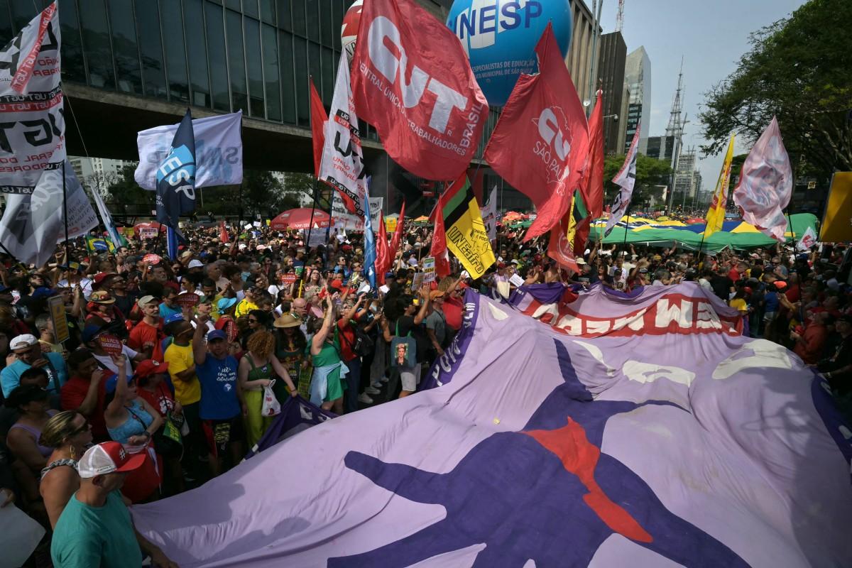 Multidão de manifestantes ocupa a Avenida Paulista, em São Paulo, durante um protesto político. Pessoas seguram uma grande faixa roxa com a silhueta de uma figura humana em vermelho e azul, além de outras faixas e bandeiras de sindicatos e movimentos sociais. Destacam-se bandeiras vermelhas da CUT (Central Única dos Trabalhadores) e da UNE (União Nacional dos Estudantes), além de uma grande bexiga azul com a sigla 