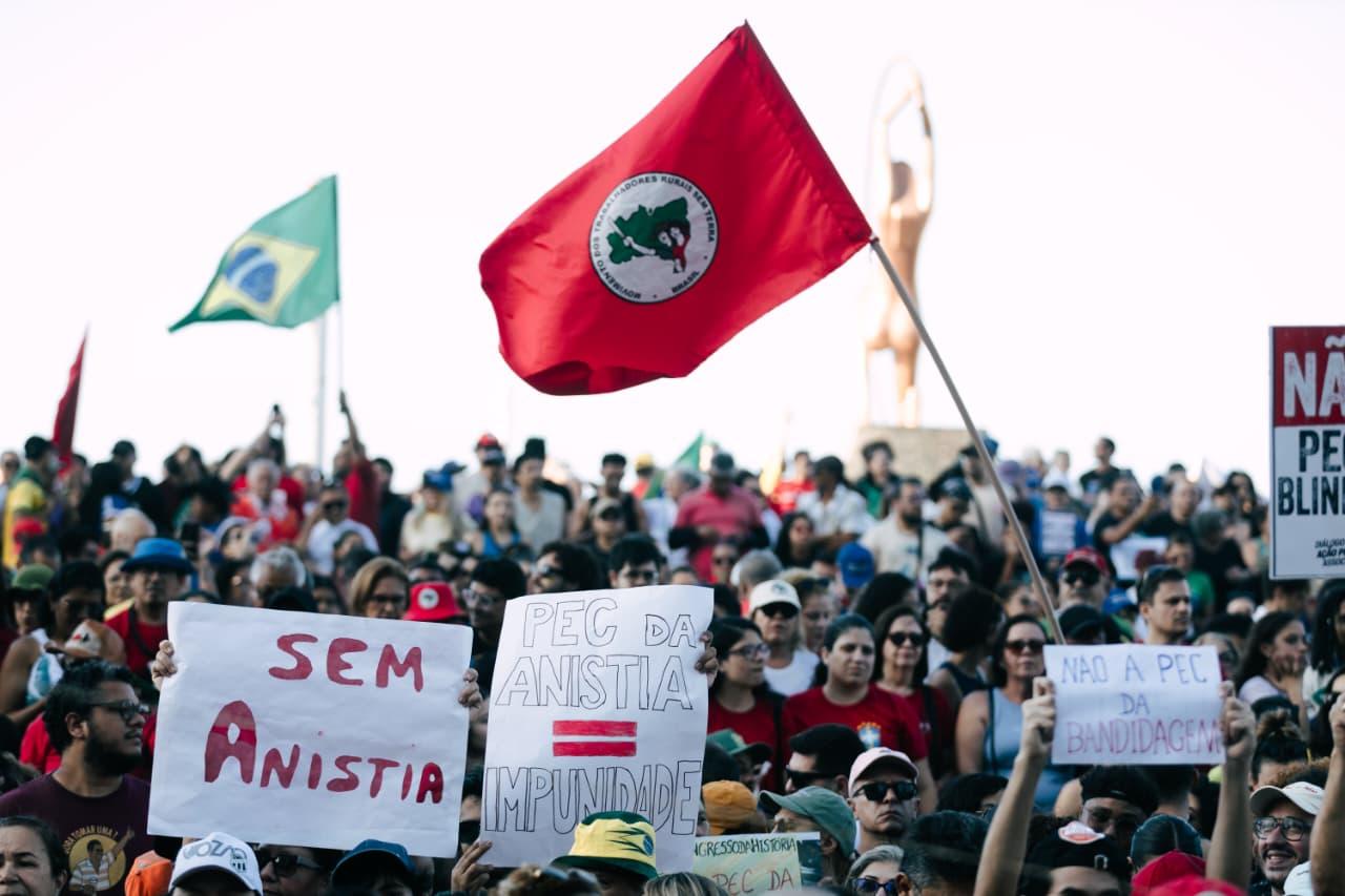 Manifestação popular com muitas pessoas reunidas, algumas segurando cartazes e bandeiras do MST. No centro da imagem, cartazes dizem 