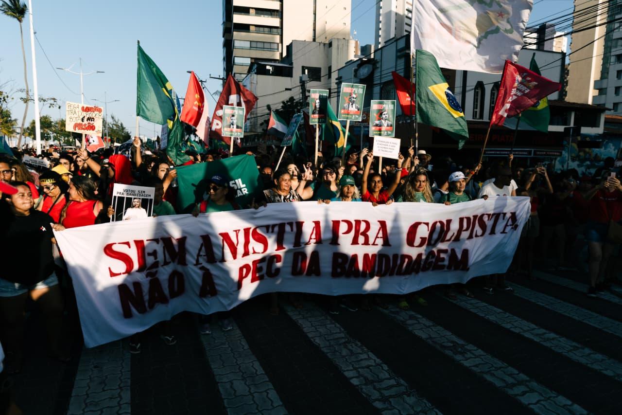 Manifestantes ocupam uma rua em Fortaleza durante protesto contra a PEC da Blindagem. Um grupo segura uma grande faixa branca com letras vermelhas onde se lê: “Sem anistia pra golpista. Não à PEC da Bandidagem”. Pessoas ao redor levantam cartazes com fotos de deputados e bandeiras do Brasil e de movimentos sociais. Ao fundo, prédios e comércio compõem a paisagem urbana.