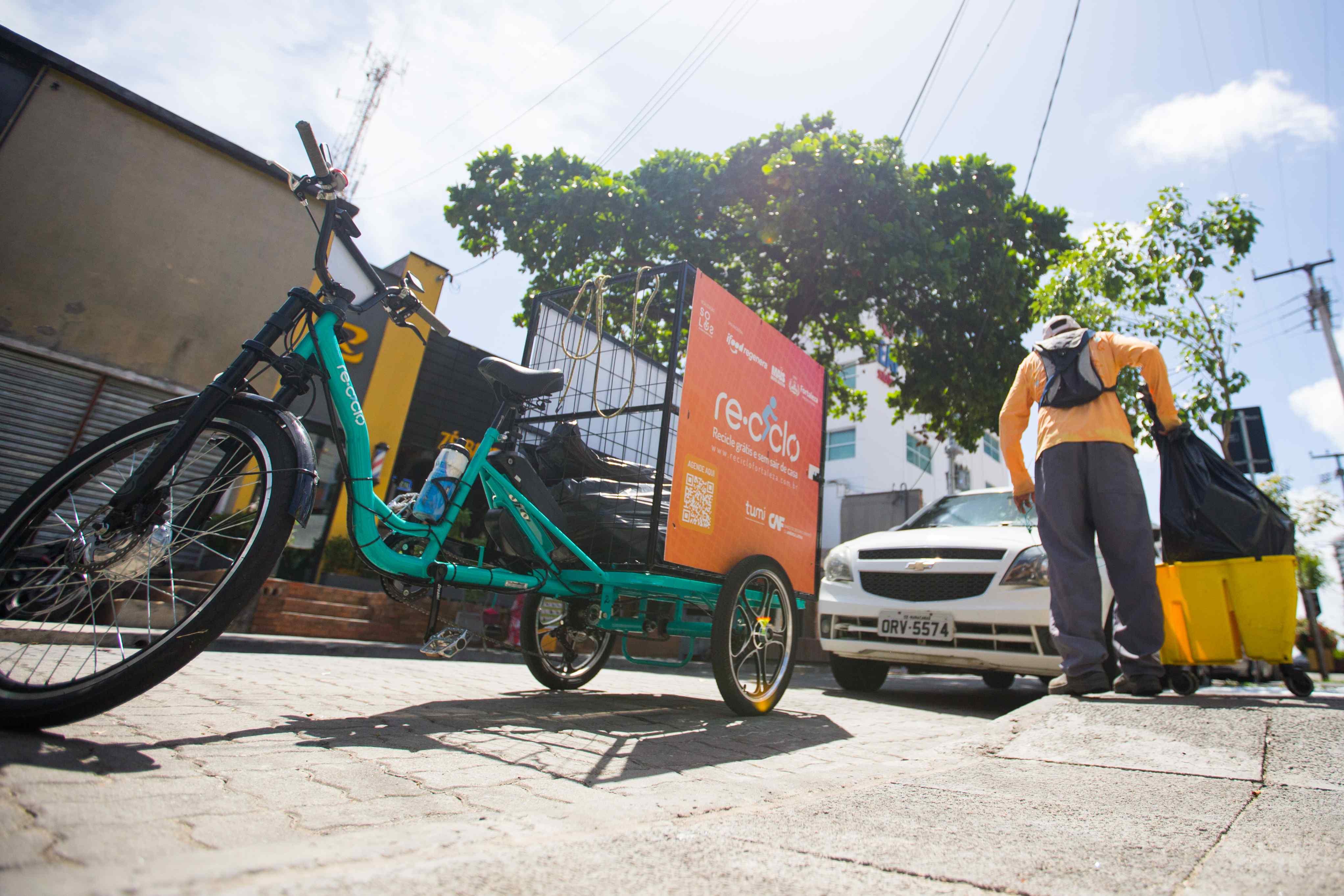 Uma pessoa usando uma camiseta laranja e calças cinza está na calçada, de costas para a câmera, carregando sacos de lixo preto. Ele está perto de um triciclo de bicicleta turquesa com um cesto de lixo preto e uma placa laranja no verso que diz 