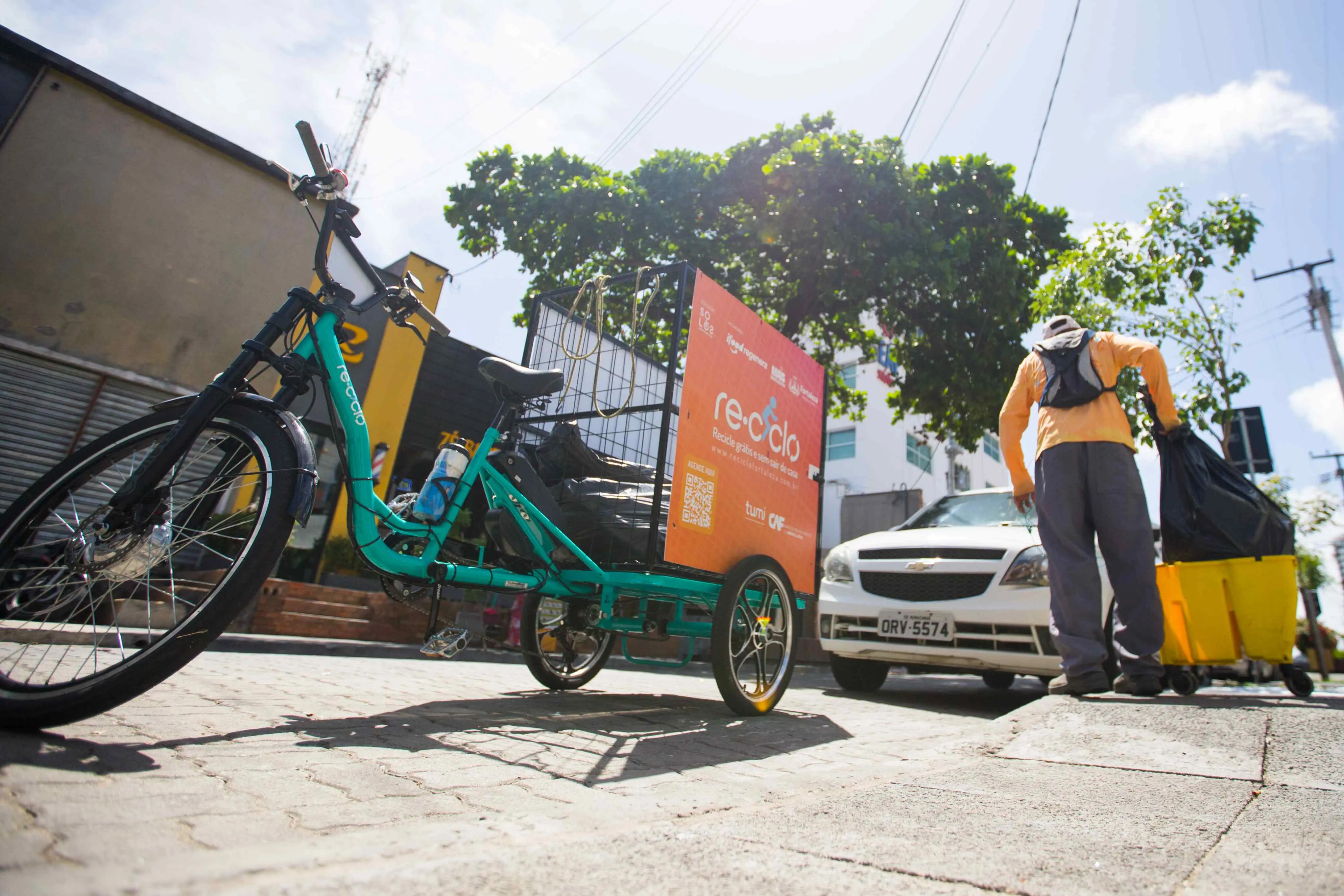 Uma pessoa usando uma camiseta laranja e calças cinza está na calçada, de costas para a câmera, carregando sacos de lixo preto. Ele está perto de um triciclo de bicicleta turquesa com um cesto de lixo preto e uma placa laranja no verso que diz 