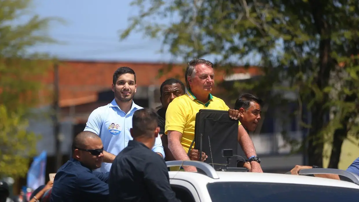 Imagem mostra o ex-presidente Jair Bolsonaro, com uma camiseta da seleção, e André Fernandes, com uma camisa azul, em cima de um carro durante carreata. Ao lado, seguranças acompanham os políticos
