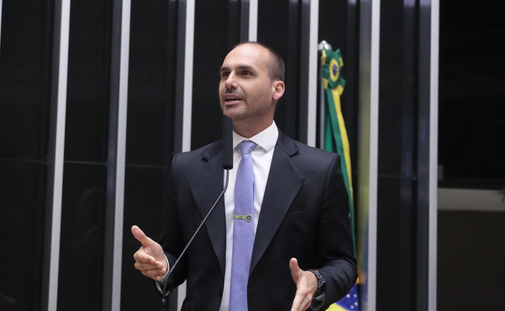 Eduardo Bolsonaro de terno preto e gravata azul faz discurso em evento público, com fundo de palco decorado, em ambiente interno de conferência ou reunião.
