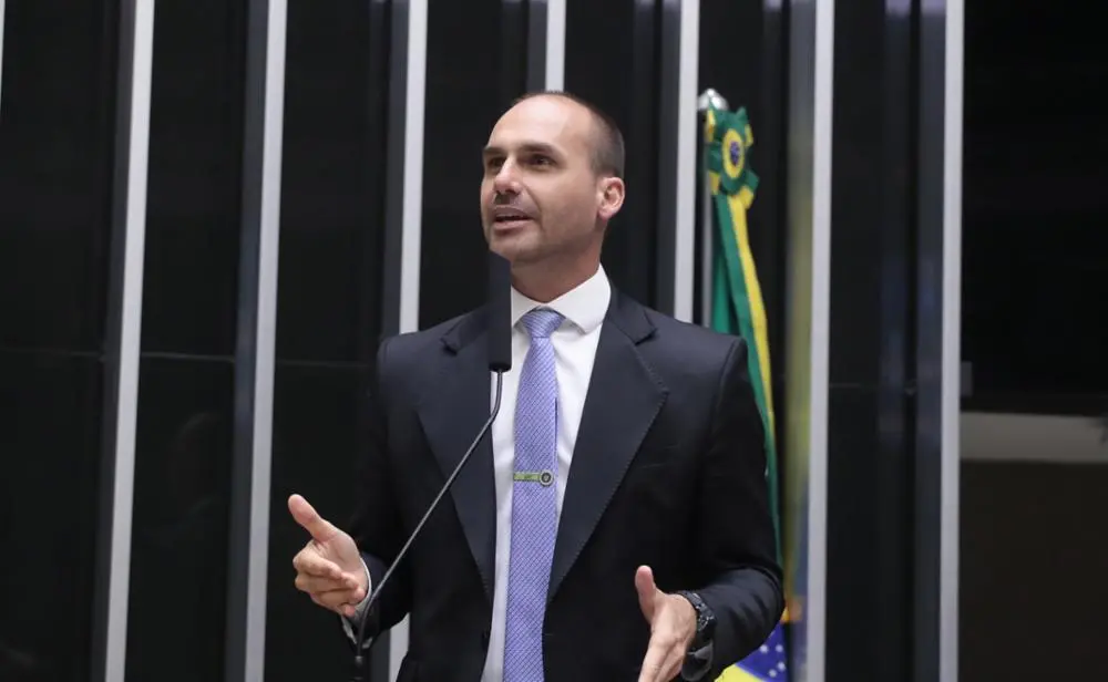 Eduardo Bolsonaro de terno preto e gravata azul faz discurso em evento público, com fundo de palco decorado, em ambiente interno de conferência ou reunião.