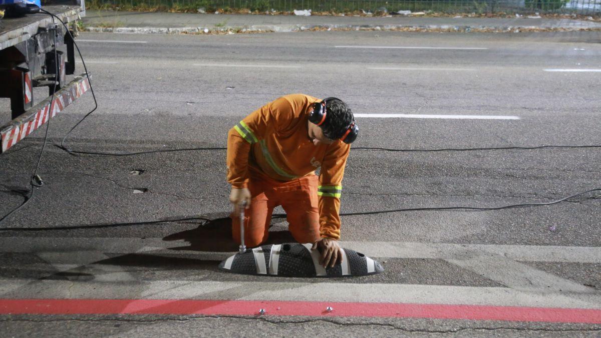 Trabalhador ajoelhado em uma via durante a instalação de uma lombada física. Ele veste uniforme laranja com faixas refletivas e protetor auricular, utilizando uma ferramenta para fixar a estrutura no chão. Há cabos e equipamentos ao redor, com parte de um veículo visível à esquerda. A cena ocorre à noite, sob iluminação artificial.