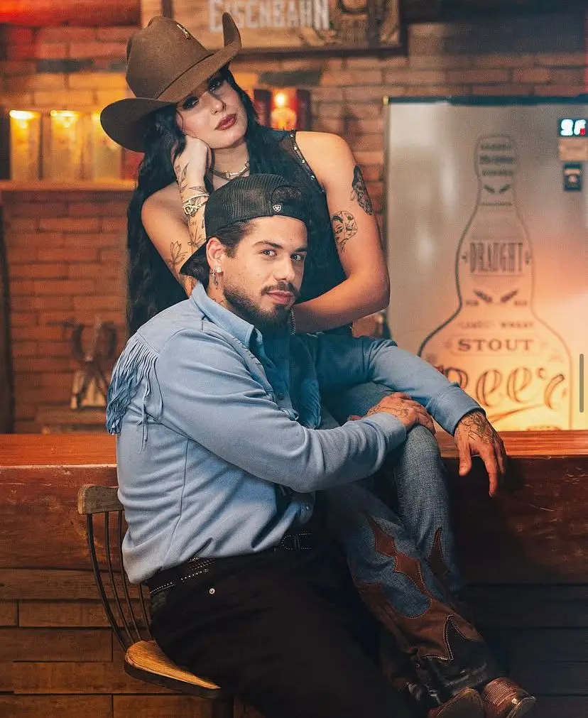 Imagem de um casal (Zé Felipe e Ana Castela) jovem e estiloso em um ambiente aconchegante, com decoração rústica, onde ela usa chapéu e tatuagens, e ele usa boné, demonstrando estilo e personalidade.