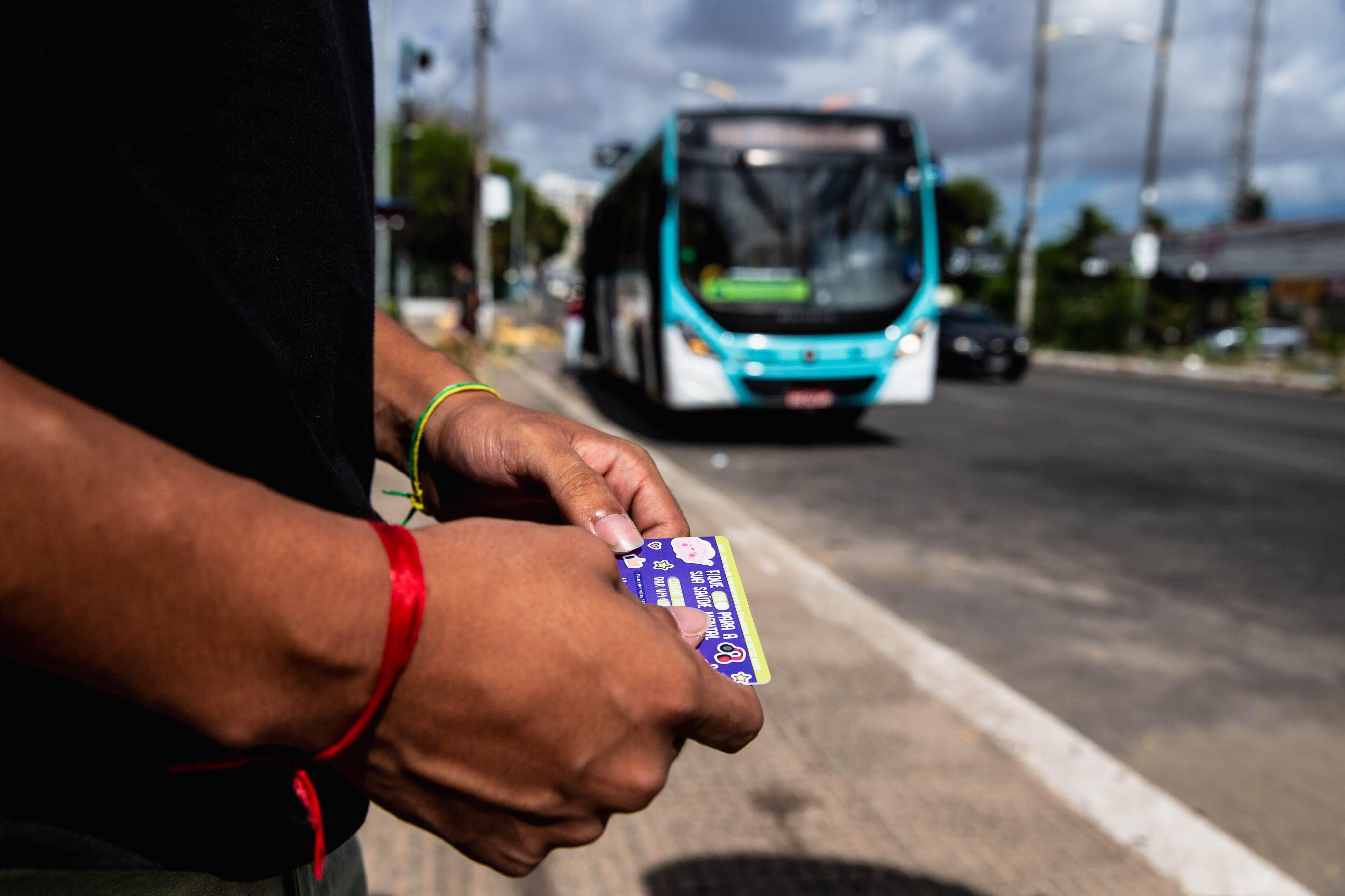 Em primeiro plano, mãos seguram um cartão de transporte, possivelmente um passe livre estudantil. Em desfoque no fundo, um ônibus azul e branco se aproxima em uma avenida sob um céu parcialmente nublado.