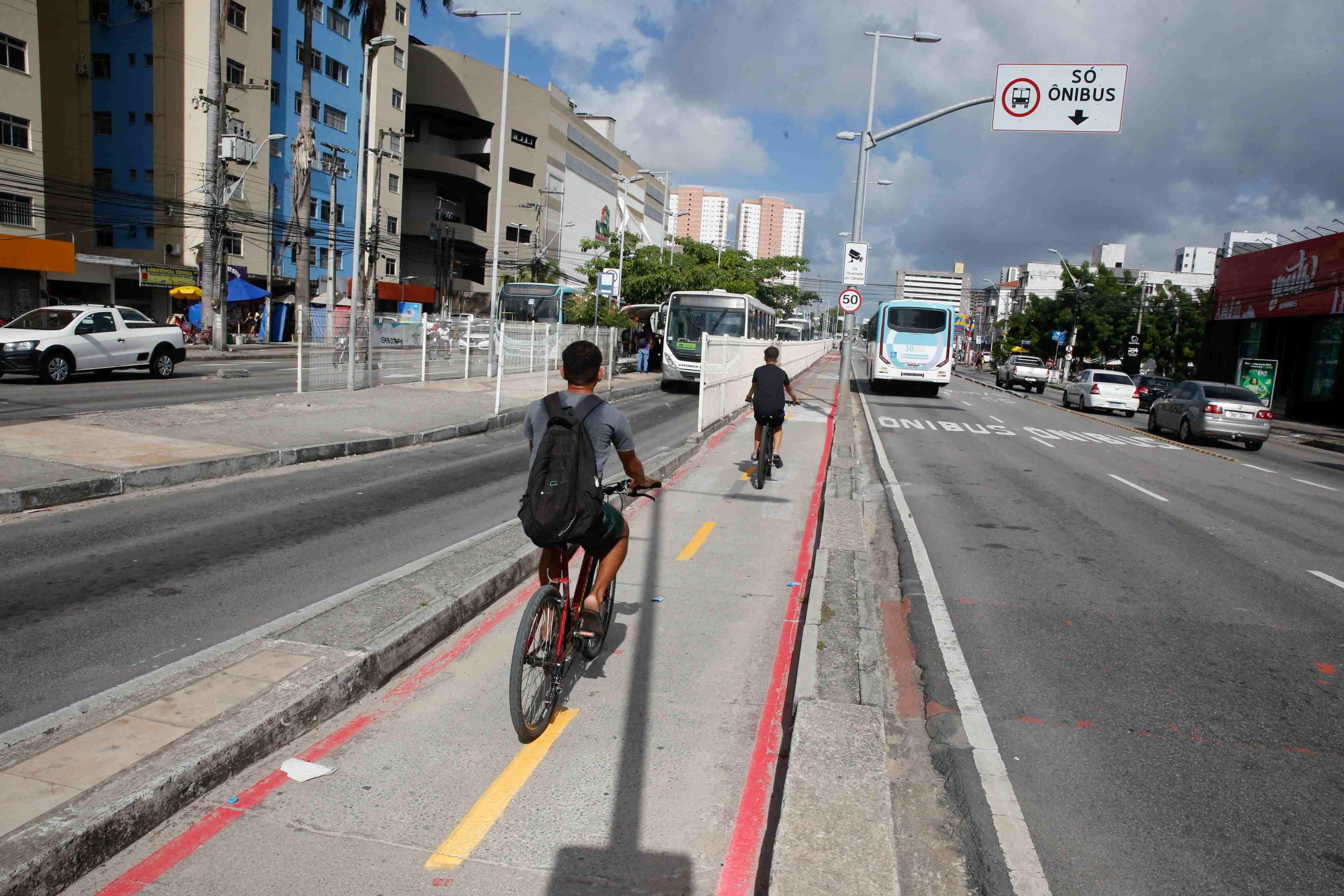 Dois ciclistas pedalam em uma ciclovia segregada por um meio-fio em uma grande avenida urbana, com o fundo repleto de prédios e ônibus de transporte público. Há uma placa visível indicando 