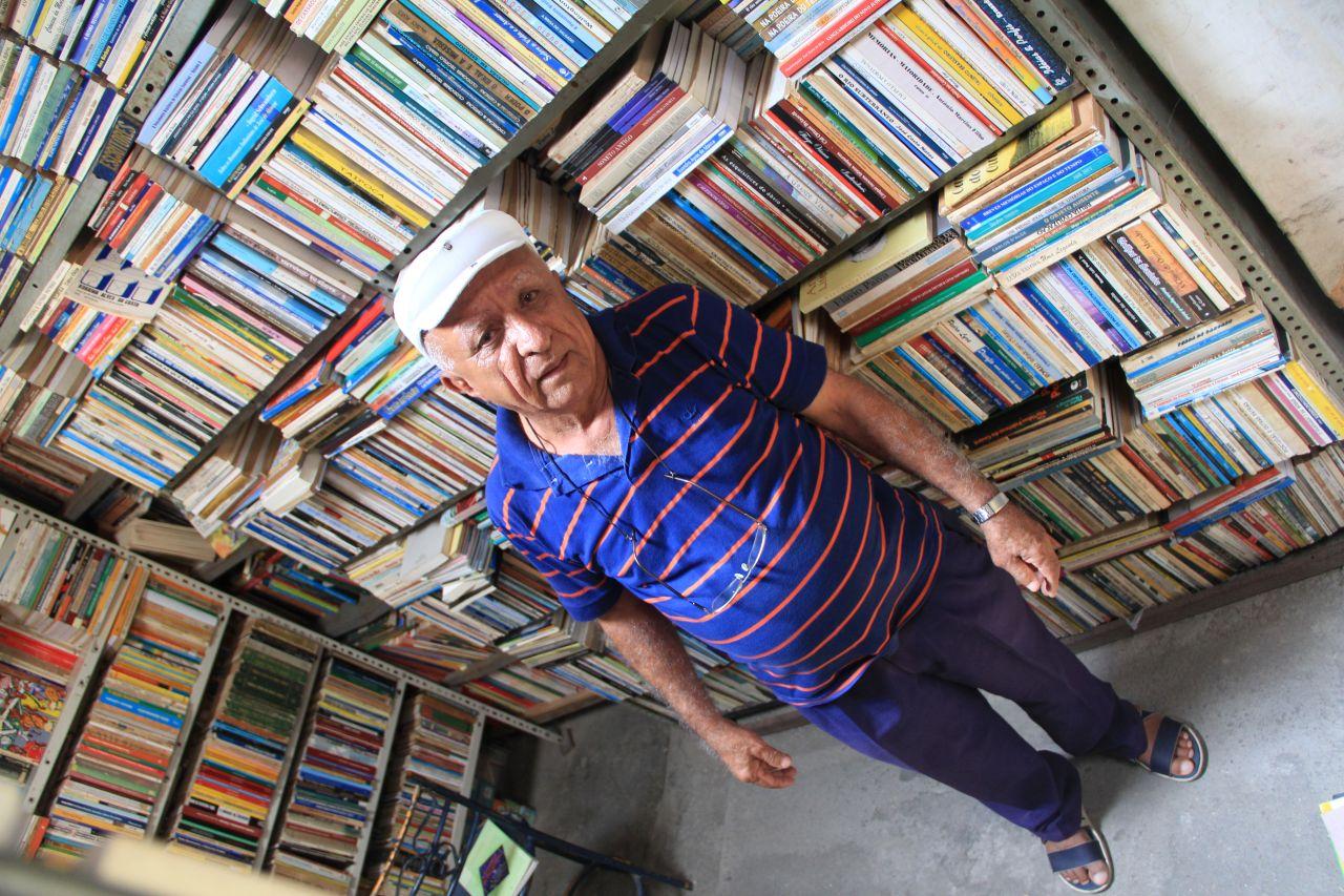 Imagem mostra Geraldo Paulo Duarte, fundador do sebo O Geraldo, um dos mais tradicionais do Centro de Fortaleza, em 2014. Com expressão série e vestindo blusa azul escura listrada de laranja, calça também em azul-escuro e boina branca, ele entre as estantes de livros do sebo.