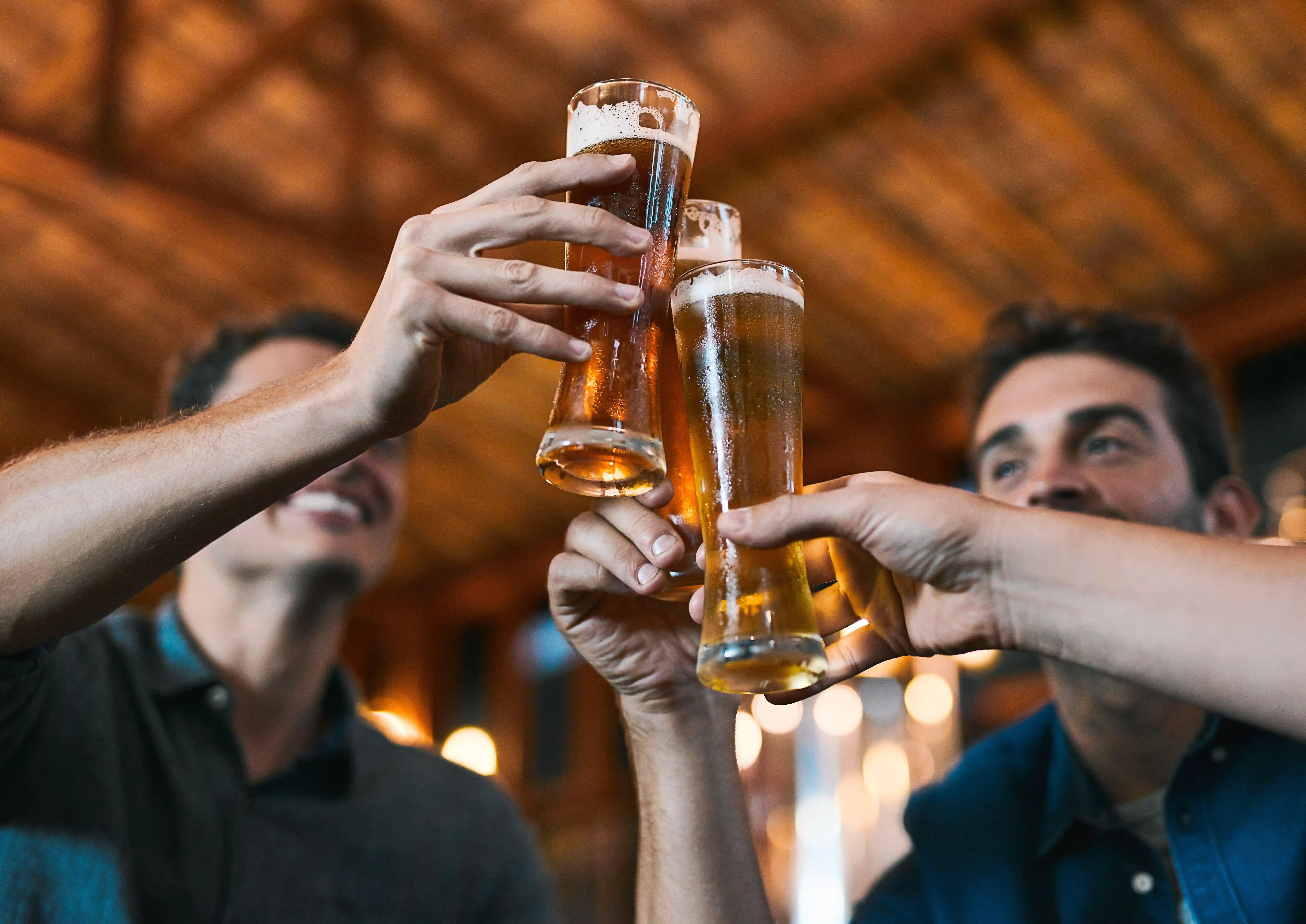 Close-up de três pessoas fazendo um brinde com copos de cerveja em um bar, em um momento de celebração. A cena social levanta o debate sobre o consumo de bebidas, como a dúvida se a cerveja sem álcool faz mal à saúde.