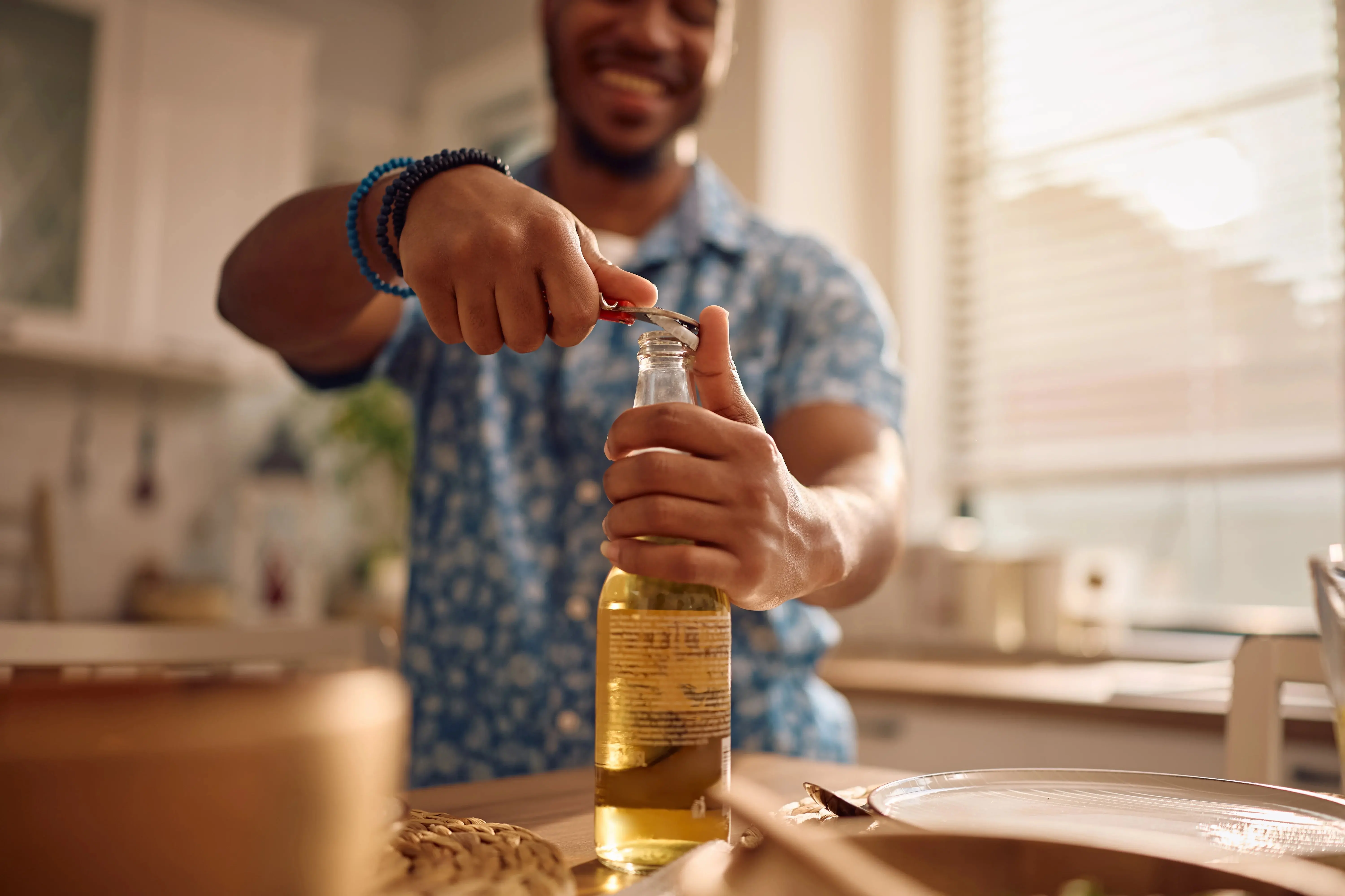 Um homem sorridente e de pele escura abre uma garrafa de cerveja clara com um abridor em uma cozinha ensolarada. A ação de consumo da bebida é o foco do debate sobre saúde, levantando a dúvida se a cerveja sem álcool faz mal à saúde