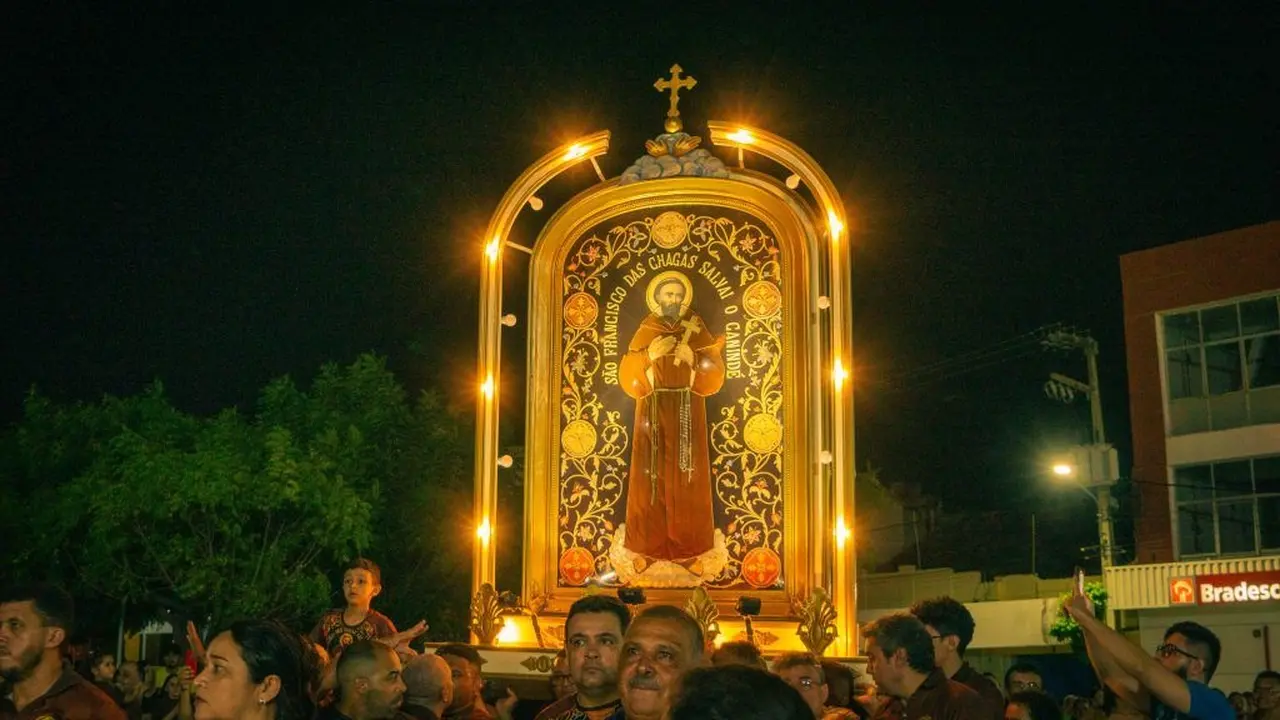 Imagem de uma procissão religiosa durante a noite, com uma imagem iluminada de São Francisco de Assis em um caminhão decorado, rodeada por pessoas participando do evento.