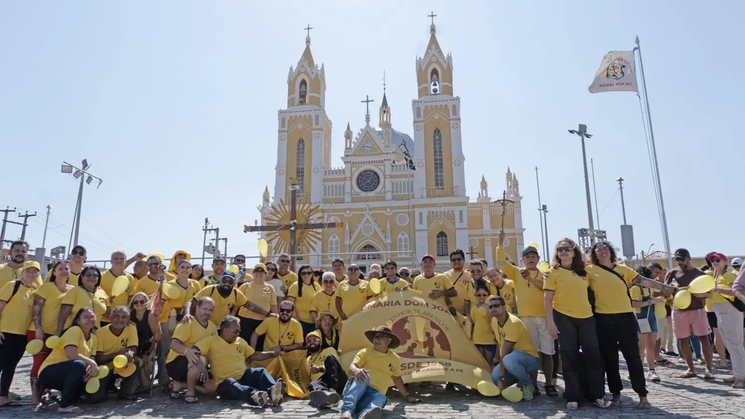 Grupo de pessoas vestidas de amarelo participando de evento religioso em frente a uma grande igreja com torres na cidade.