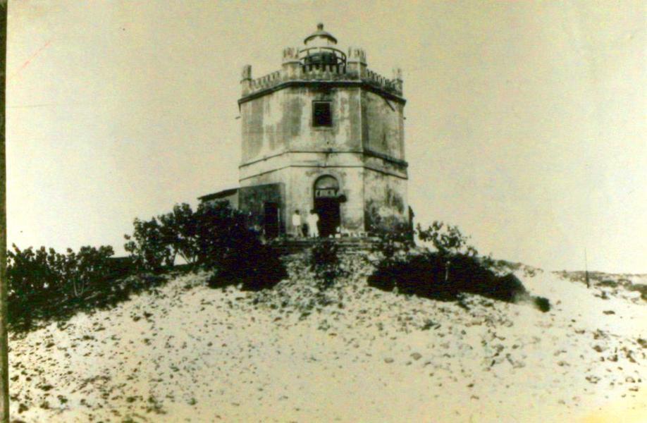 Fotografia em preto e branco de 1918 mostrando o antigo Farol do Mucuripe, em Fortaleza. A construção tem formato octogonal, com janelas e detalhes arquitetônicos na parte superior, coroada por uma lanterna no topo. O farol está erguido sobre uma duna de areia, rodeado por vegetação rasteira, e algumas pessoas aparecem na entrada da edificação.
