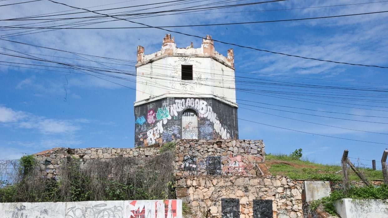 Farol antigo do Mucuripe em Fortaleza. A construção octogonal aparece desgastada, com paredes pichadas e sem cobertura, cercada por fios elétricos no céu azul. A base de pedra também exibe pichações e sinais de abandono, com vegetação crescendo ao redor.
