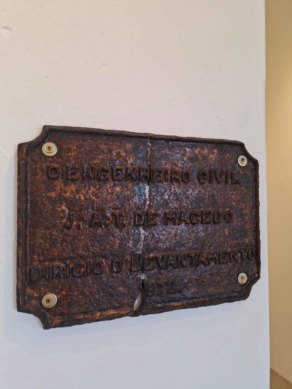 Placa metálica antiga, enferrujada e fixada em uma parede branca, com inscrições em relevo parcialmente desgastadas. O texto visível diz: “O ENGENHEIRO CIVIL J. A. T. DE MACEDO DIRIGIO O LEVANTAMENTO 1872”.