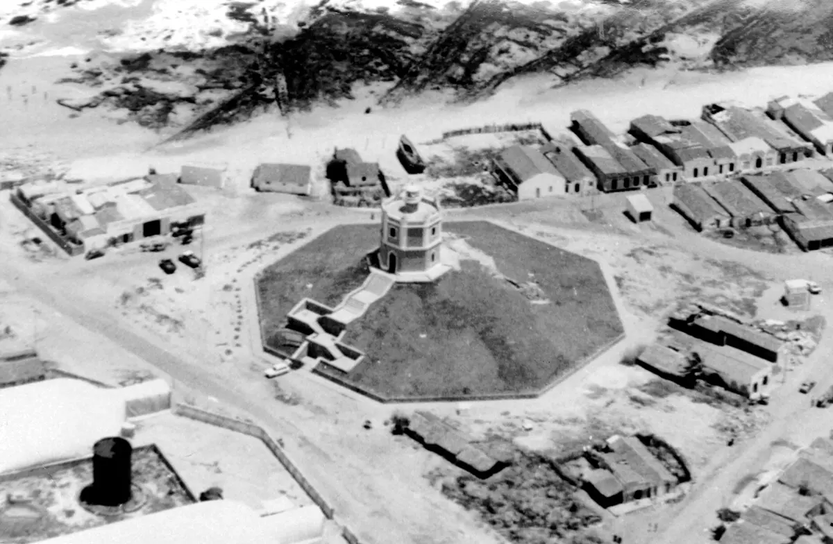 Fotografia aérea em preto e branco da década de 1940 mostrando o Farol Velho do Mucuripe, em Fortaleza. A construção octogonal com escadaria e cercada por um muro aparece no centro da imagem, em meio a uma área de casas simples e ruas de terra próximas à praia.