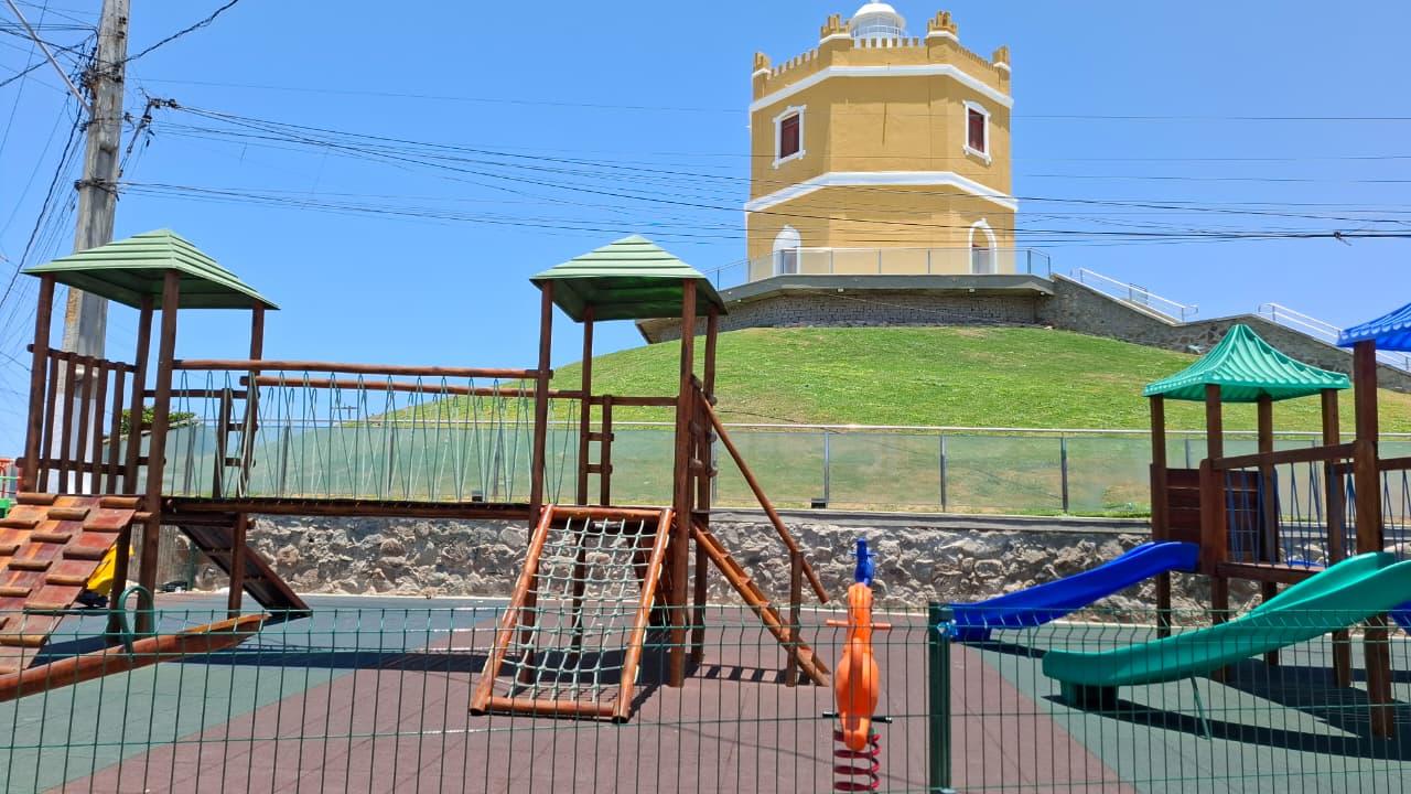 Área de brinquedopraça com escorregadores e estruturas de madeira ao ar livre, em frente ao Farol Velho do Mucuripe restaurado, pintado em amarelo e branco, localizado sobre uma colina gramada sob céu azul.