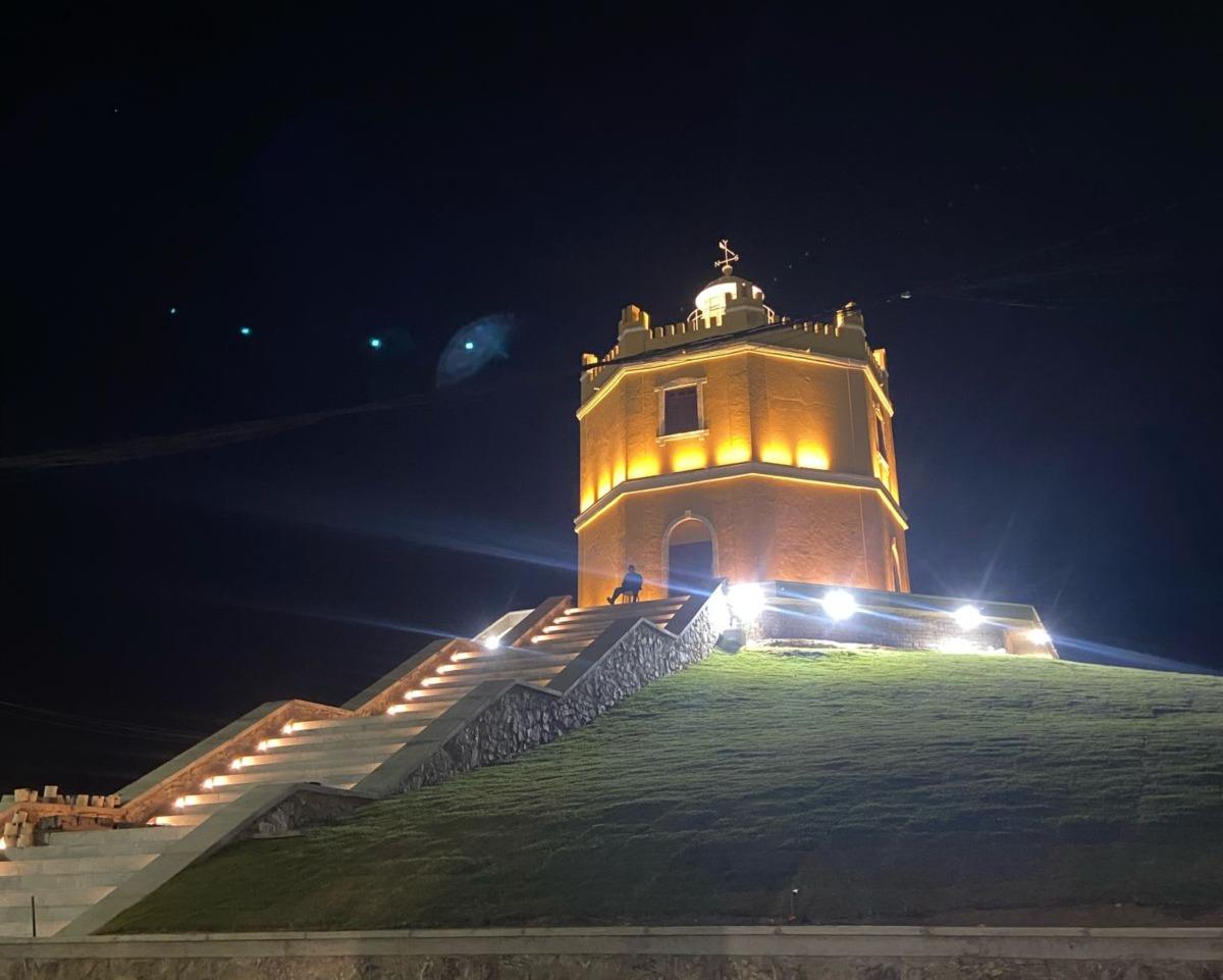 Farol Velho do Mucuripe iluminado à noite com luzes amarelas e brancas, visto de baixo do morro gramado com escadaria de pedra e degraus iluminados que levam até a entrada do farol.