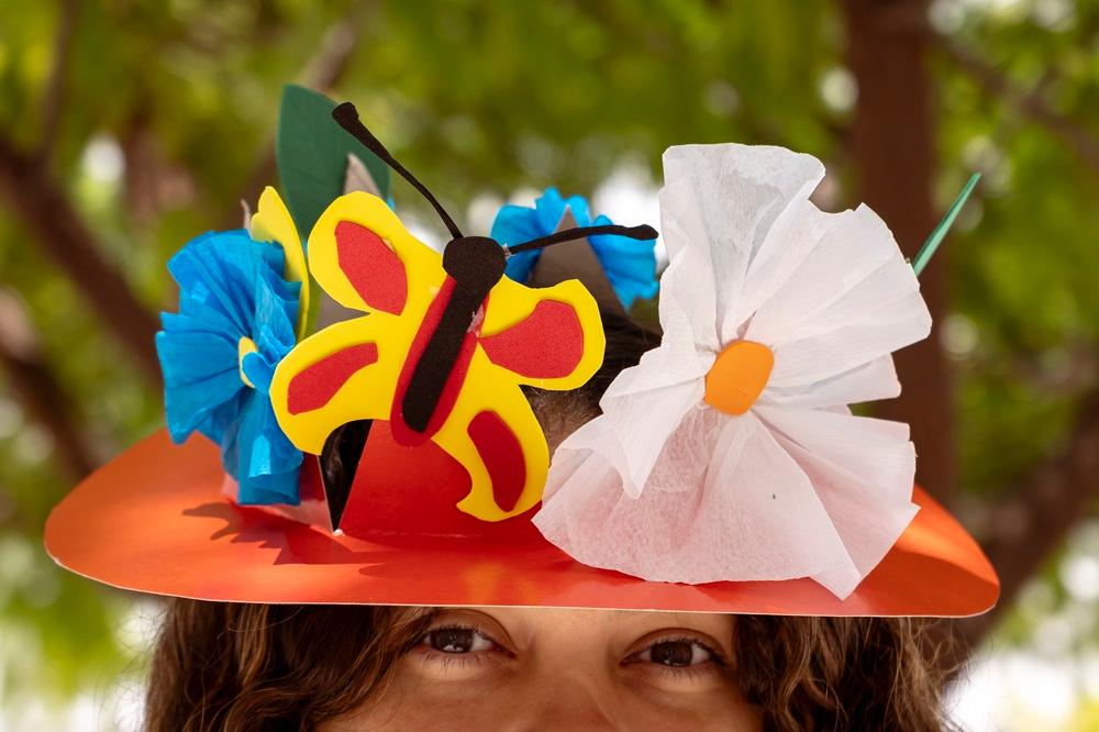 Na imagem, mulher veste chapéu colorido e criativo durante oficina do Ateliê Criança Cria Chapéu Divertido, promovido pela Pinacoteca do Ceará em alusão ao Dia da Criança