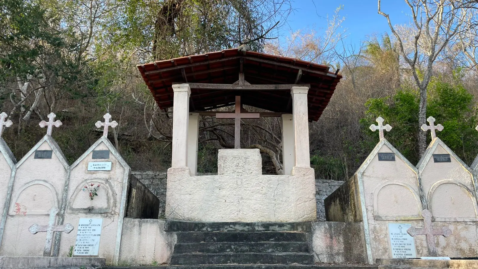 Cemitério com muro de cimento, grade de ferro e cruzes brancas em um ambiente ao ar livre, cercado por árvores e vegetação, sob céu azul.